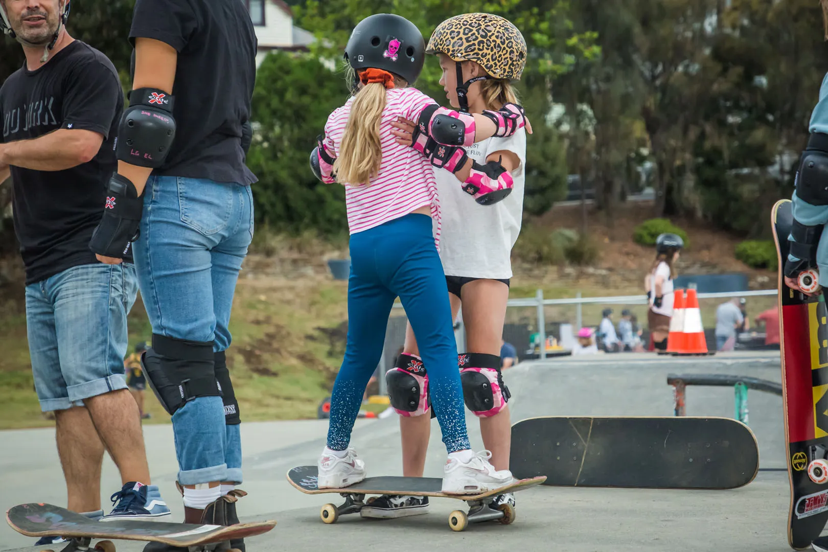 Two young skaters in helmets and pads hugging at the skatepark, boards at their feet